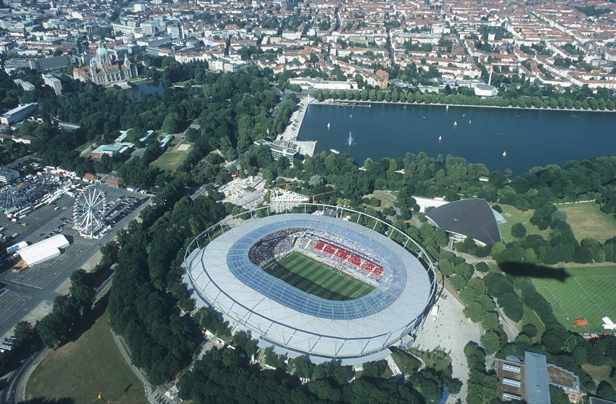 O estádio cercado de verde entre o lago Maschsee e a prefeitura nova, com a arquibancada cheia em dia de jogo.