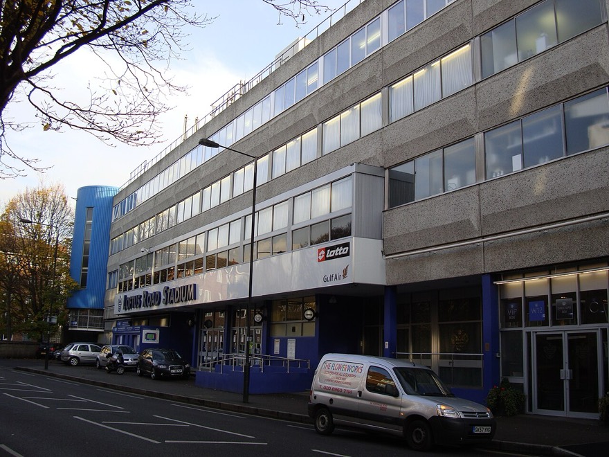 A entrada da South Africa Road, com o letreiro de Loftus Road na fachada e os escritórios do clube em cima da bilheteria.
